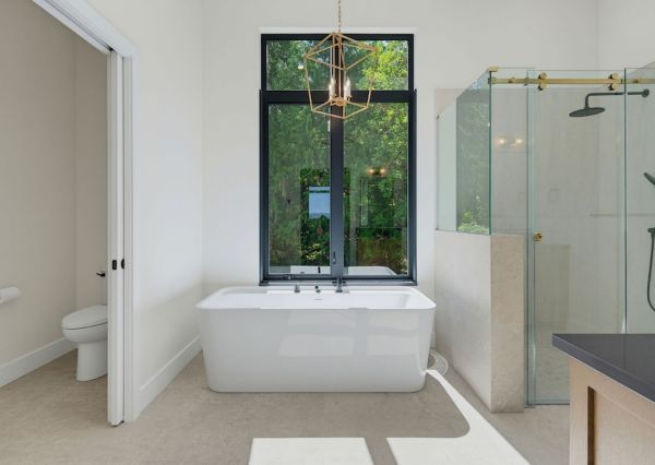 Primary ensuite with freestanding tub, black-framed window, glass shower and brass pendant light.
