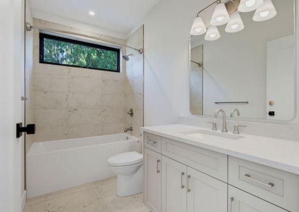 Bright secondary bathroom with white vanity, stone-look tile, bathtub and black-framed window.