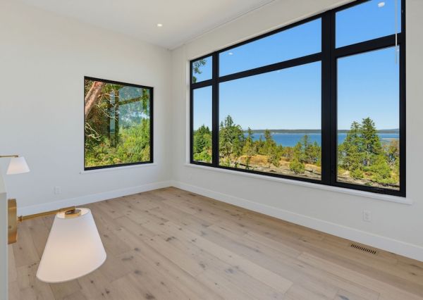 Bright waterfront bedroom with large black-framed windows overlooking the ocean and surrounding trees.